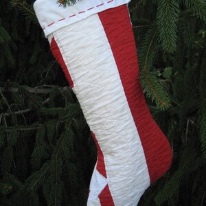 May include: A handmade Christmas stocking with a white top and a red and white striped design. The stocking features a white star and is hanging in front of a green evergreen tree.
