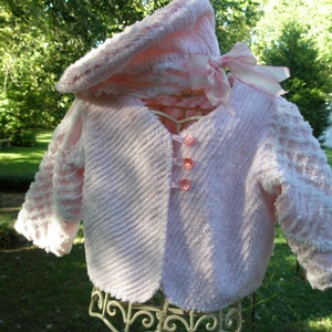 May include: A pink baby jacket with a hood and a bow. The jacket has a ruffled trim and is made of a soft, fluffy material.