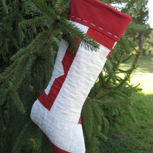 May include: A handmade Christmas stocking in red and white hangs from a tree branch. The stocking features a red cuff and toe, with a white body accented by red geometric shapes. The design is quilted, and the stocking is suspended by a red loop.