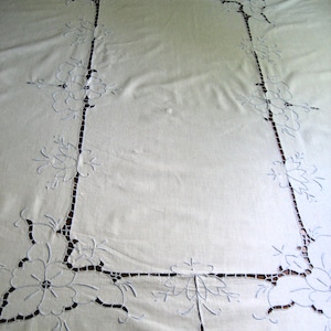 May include: A rectangular white tablecloth featuring intricate cutwork embroidery along the border. The design includes floral motifs and openwork patterns that create a decorative frame around the solid center of the fabric.