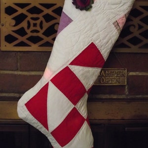 May include: A quilted Christmas stocking featuring a geometric patchwork pattern in white and red, with accents of pink and purple. A dark red and green fabric flower is attached to the top cuff of the stocking, which is constructed from stitched fabric.