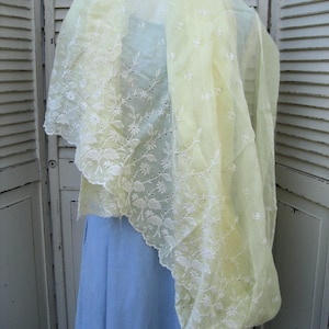 May include: A sheer, pale yellow shawl with intricate white floral embroidery along the edges. The delicate fabric drapes over a light blue garment, creating a soft, layered effect. The shawl's design features a scalloped edge.