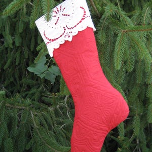 May include: A quilted red Christmas stocking with a white lace cuff, hanging from a tree branch. The stocking has a detailed pattern and a scalloped edge on the cuff. The background is a green evergreen tree.