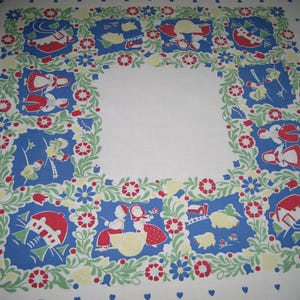 May include: A white square tablecloth featuring a central blank area surrounded by a border of blue squares. Each blue square contains illustrations of figures, houses, and farm scenes in red, yellow, and green, connected by floral vines.