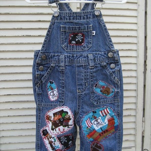 May include: Blue denim overalls with various colorful patches featuring pirate ships, skulls, and treasure chests. The overalls have adjustable straps and two front pockets.