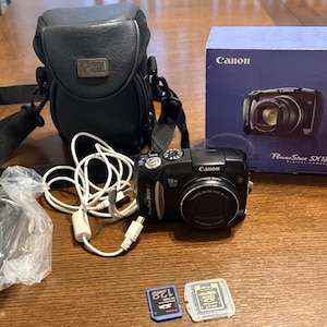 May include: A black Canon PowerShot SX120 IS digital camera with a 10x zoom lens, a black carrying case, a white USB cable, a blue 128MB SD memory card, a clear plastic SD card case, and the original blue Canon product box with camera imagery.