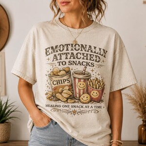 Op de afbeelding: Beige t-shirt met de tekst "Emotionally Attached to Snacks" en illustraties van chips, friet en een drankje. Het shirt heeft een vintage design met een distressed look, en de tekst "Healing one snack at a time" staat er ook op.