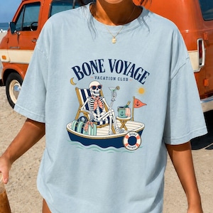 May include: Light blue t-shirt with the text "BONE VOYAGE VACATION CLUB" and a graphic of a skeleton in sunglasses relaxing on a boat, holding a cocktail. The shirt is a chambray color.