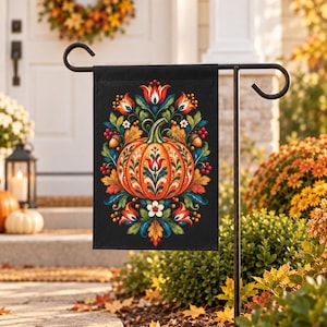 May include: A rectangular garden flag hanging on a black metal stand features a central orange pumpkin decorated with intricate folk-art style floral patterns in red, blue, and green, surrounded by autumn leaves, acorns, and berries on a black background.