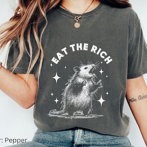 Puede incluir: Una camiseta gris oscuro con un gráfico blanco de una zarigüeya con el texto "Eat the Rich" encima. La zarigüeya está de pie sobre sus patas traseras con la boca abierta y estrellas a su alrededor.