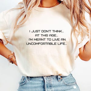May include: A white t-shirt with the text "I just don't think... at this age... I'm meant to live an uncomfortable life..." printed on it. The shirt is being worn by a person with blonde hair and blue jeans.