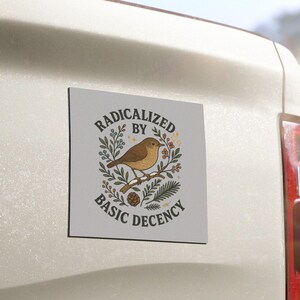 May include: A square magnet with a light grey background. It features a brown bird on a branch, encircled by leaves and berries. The text "RADICALIZED BY BASIC DECENCY" is arranged around the bird in a circular design.