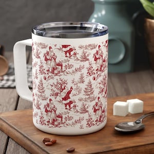 Op de afbeelding: Een witte reismok met een rood en wit toile de Jouy-patroon met kerstmanfiguren en winterse taferelen. De mok heeft een zilveren rand en een wit handvat. De mok staat op een houten snijplank.