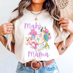 May include: Ivory-colored t-shirt with the words "Mahi Mama" in lavender script. The shirt features a colorful illustration of a peacock, flowers, and Mahjong tiles. The shirt is paired with denim shorts and a brown belt.