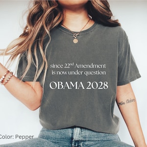 May include: Pepper-colored t-shirt with the text "since 22nd Amendment is now under question OBAMA 2028." The shirt is paired with blue jeans and white sneakers. The color of the shirt is labeled as "Pepper."