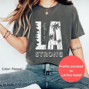 以下が含まれることがあります： 白い文字で「LA」と「Los Angeles」「STRONG」が書かれたグレーのTシャツ。ヤシの木のグラフィックが描かれている。また、「Profits donated to LA Fire Relief」というテキストも見える。