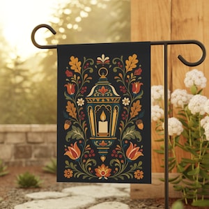 May include: A black garden flag with a lantern design and a lit candle. The lantern is surrounded by colourful floral and leaf patterns in orange, red, yellow, and green. The flag is displayed on a black metal stand in a garden setting.