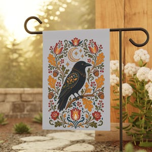 Peut inclure: Un drapeau de jardin décoratif avec un motif de corbeau noir perché sur une branche, entouré de motifs floraux et de glands. Le design comprend un croissant de lune et des étoiles, avec des accents rouges, orange et jaunes. Le drapeau est sur un support en métal noir.