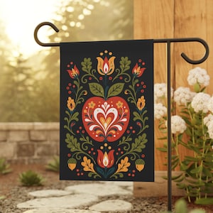 Pode incluir: Bandeira de jardim preta com um design de arte popular. Apresenta um coração de maçã vermelha rodeado por motivos florais e de folhas laranja, amarelas e verdes. A bandeira é exibida em um suporte de metal preto, adicionando um toque de charme rústico ao jardim.