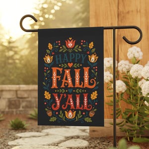 May include: Black garden flag with the words "HAPPY FALL Y'ALL" in orange and white lettering. The flag is decorated with floral and leaf designs in red, yellow, and blue. The flag is displayed on a black metal stand in a garden setting.