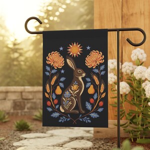 Puede incluir: Una bandera de jardín rectangular con fondo oscuro y una ilustración central de un conejo marrón. El conejo está rodeado de crisantemos naranjas, espigas de trigo, peras y bayas rojas. Un sol y estrellas se sitúan sobre el conejo.