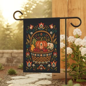 Op de afbeelding: Zwarte tuinvaandel met een mand vol herfstproducten, waaronder een grote oranje pompoen, peren en appels. De mand is omgeven door bloemen- en bladmotieven in rood, teal en wit. De vlag staat op een zwarte metalen standaard.