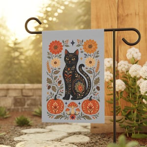 May include: A rectangular garden flag featuring a black cat illustration centred among orange and yellow flowers, two orange pumpkins, and small mushrooms. The design includes intricate folk-style patterns in orange, red, and green on a light grey background.
