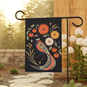 Op de afbeelding: Een rechthoekige tuinvlag met een zwarte achtergrond met een centrale fazantillustratie omringd door oranje kaki's, tarwestengels en drie grote gestileerde bloemen in tinten oranje, geel en wit met kleine steraccenten.