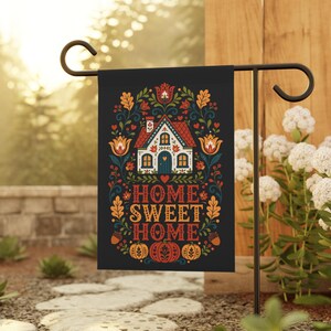 Op de afbeelding: Een zwarte tuin vlag met een centrale illustratie van een huis met een rood dak, omringd door kleurrijke bloemen- en bladmotieven. De tekst HOME SWEET HOME staat in het midden gedrukt, met pompoenen en eikels onderaan het ontwerp.