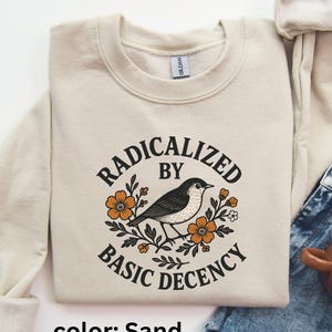 Könnte beinhalten: Sandfarbenes Sweatshirt mit dem schwarzen Text "RADICALIZED BY BASIC DECENCY", der eine Vogelillustration mit orangefarbenen Blumen umgibt. Das Sweatshirt ist gefaltet, mit einer Jeans und einer Kaffeetasse im Hintergrund.
