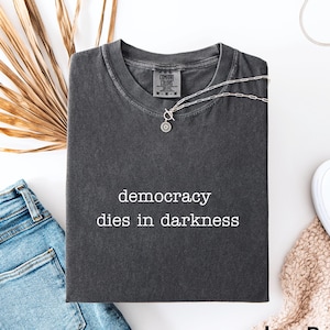 Op de afbeelding: Een donkergrijs t-shirt met de witte tekst "democracy dies in darkness". Het shirt wordt gecombineerd met een blauwe jeans, een zilveren ketting en witte instappers. De kleur van het shirt is "Pepper".