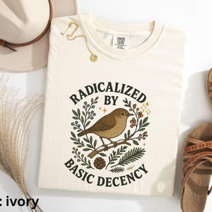 以下が含まれることがあります： 「RADICALIZED BY BASIC DECENCY」の文字と鳥のイラストが描かれたアイボリー色のTシャツ。シャツは折りたたまれており、近くに帽子、サンダル、ジュエリーが見えます。シャツは柔らかい素材で作られています。