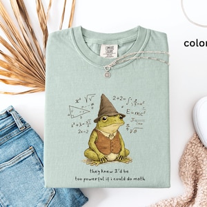 May include: A light green t-shirt featuring a cartoon frog in a wizard hat and vest, with math equations around it. The text reads "they knew I'd be too powerful if I could do math". The shirt is displayed with blue jeans and white shoes.
