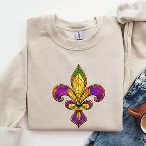 Op de afbeelding: Beige crewneck sweatshirt met een kleurrijk glas-in-lood fleur-de-lis ontwerp. De fleur-de-lis is in paars, geel, groen en oranje. De sweatshirt wordt getoond met een spijkerbroek en een koffiemok.