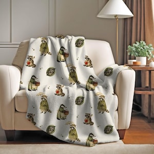 Op de afbeelding: Een witte deken met een herhalend patroon van wasberen in groene jassen en hoeden, met manden en paraplu's. De deken is over een crèmekleurige fauteuil gedrapeerd. Het patroon bevat ook groene bladeren en rode bessen.