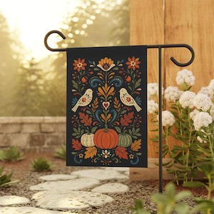 Könnte beinhalten: Eine schwarze Gartenflagge mit einem herbstlichen Design. Das Design umfasst zwei weiße Vögel, Kürbisse und farbenfrohe Blumen- und Blättermuster. Die Flagge wird auf einem schwarzen Metallständer in einem Garten gezeigt.