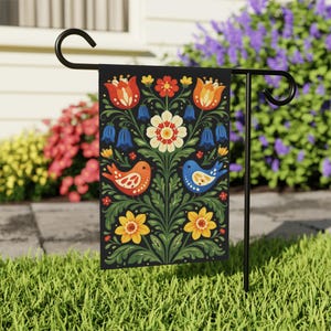 Può includere: Una bandiera da giardino nera con un colorato motivo floreale e di uccelli. Il design include tulipani arancioni, fiori gialli e bianchi, fiori blu a forma di campana e due uccelli. La bandiera è esposta su un supporto in metallo nero.