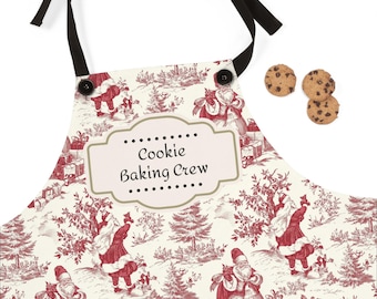 May include: A cream-colored apron with a red Christmas-themed toile pattern featuring Santa Claus figures, gifts, and trees. The apron has black ties and the text "Cookie Baking Crew" on the front. Three chocolate chip cookies are next to the apron.