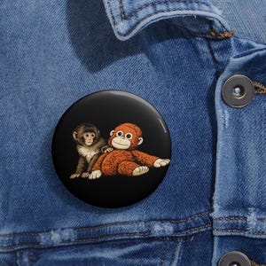 May include: A round black button featuring two cartoon monkeys. One monkey is brown and white, the other is orange. The button is set against a blue denim background.