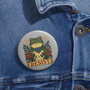 May include: A round, light gray button featuring a cartoon frog with a blue bandana, surrounded by roses and leaves. The word "RESIST" is printed on a banner below the frog, with lightning bolt accents.