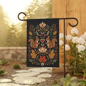 May include: A black garden flag featuring a symmetrical design with two brown squirrels facing each other, a red heart, and floral elements. The design includes orange, yellow, and green leaves, berries, and flowers. The flag is displayed on a black metal stand.