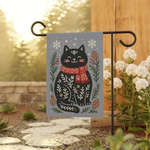 May include: A gray garden flag featuring a black cat wearing a red patterned scarf. The cat is surrounded by green leaves, berries, snowflakes, and pinecones. The flag is displayed on a black metal garden stake.