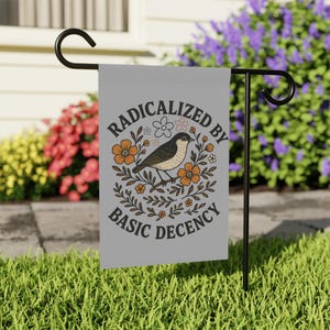 Peut inclure: Un drapeau de jardin gris avec le texte "RADICALIZED BY BASIC DECENCY" et une illustration d'oiseau entourée de fleurs. Le drapeau est suspendu à un support en métal noir dans un jardin avec de l'herbe verte et des fleurs colorées.