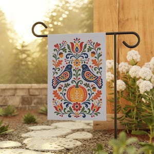 May include: A garden flag with a folk art design. It features two blue birds, a pumpkin, and floral elements in orange, red, and blue. The flag is displayed on a black metal stand in a garden.