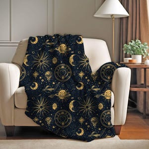 Peut inclure: Une couverture bleu marine avec un motif céleste doré de soleils, lunes, planètes et étoiles. La couverture est drapée sur un fauteuil crème, créant un accent décoratif et confortable pour un espace de vie.