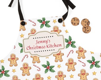 Op de afbeelding: Witte schort met een patroon van peperkoekmannen, zuurstokken, hulst en sterren. De schort heeft zwarte banden en knopen. De tekst "Jenny's Christmas Kitchen" staat erop. Drie chocolate chip cookies zijn ook zichtbaar.