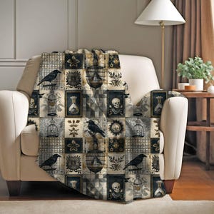 Op de afbeelding: Een decoratieve sprei met patroon op een crèmekleurige fauteuil. De sprei heeft een patchwork-ontwerp met zwarte raven, vogelkooien, schedels en bloemmotieven in een gedempt kleurenpalet van zwart, beige en grijs. De sprei voegt een gotische touch toe aan de kamer.