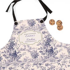 May include: A white apron with a blue toile pattern featuring a landscape scene. The apron has a black adjustable neck strap and two black buttons. A gold label with the text "Emma's Kitchen" is centered on the apron.