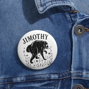 Op de afbeelding: Een ronde witte button met een zwarte inktillustratie van een wasbeer die door een veld met wilde bloemen loopt. De tekst JIMOTHY is in een vetgedrukt, zwart schreeflettertype in een boog boven de wasbeerfiguur gedrukt.