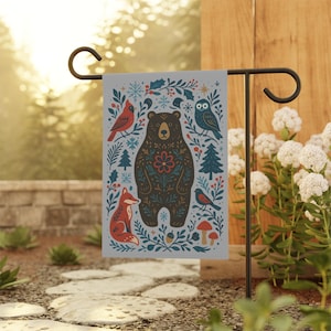 Puede incluir: Bandera de jardín decorativa con un diseño de bosque. Presenta un oso pardo, un zorro rojo, un búho, un cardenal y otros elementos del bosque. La bandera se muestra en un soporte de metal negro, añadiendo un toque de naturaleza a cualquier jardín.